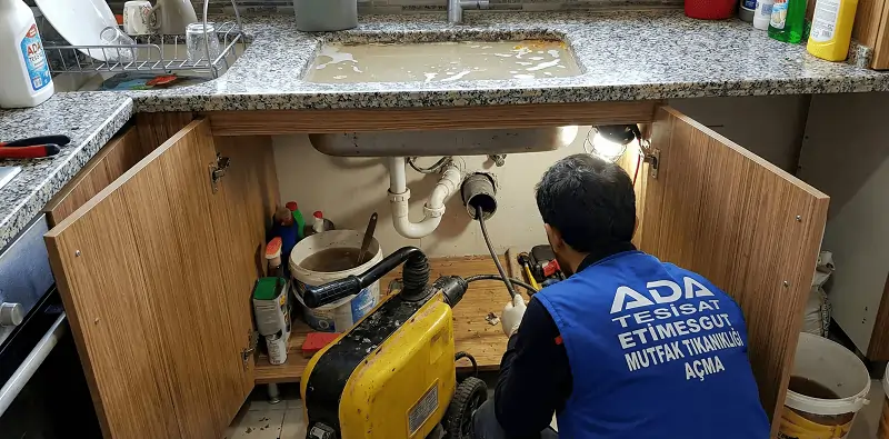 Etimesgut'ta tıkanmış ve su dolu mutfak lavabosu alt dolabında, pimaş gider borusuna sarı renkli robot makine ile müdahale eden Ada Tesisat ustası.