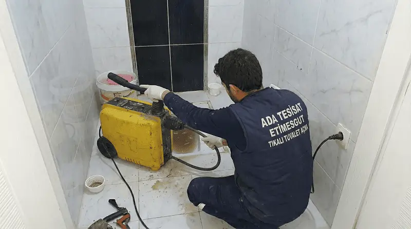 Etimesgut'ta bir Ada Tesisat ustası, tıkanmış alaturka tuvaleti kırmadan, sarı renkli robot kanal açma makinesi kullanarak açarken.